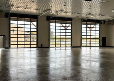 Inside a luxury country man cave showing three large glass overhead garage doors reflecting light onto a glossy finished shop floor.